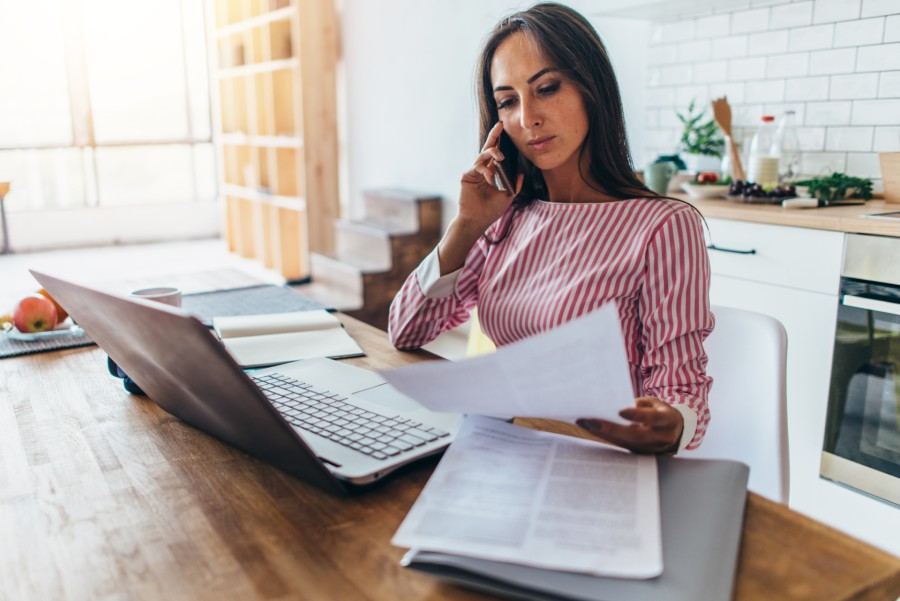 mulher ao telefone, segurando documento em frente ao notebook negociando