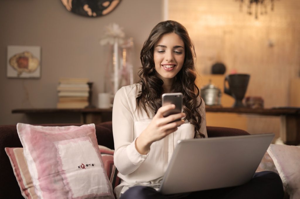 Mulher jovem sorrindo ao olhar celular com notebook aberto à frente