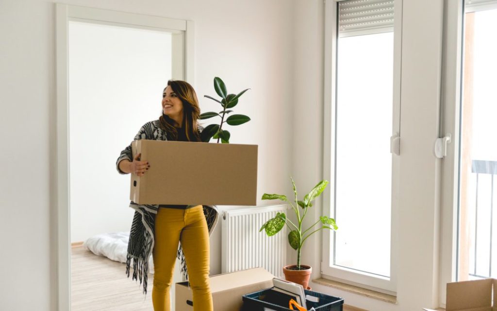 Mulher sorrindo entrando em apartamento segurando caixa de papelão